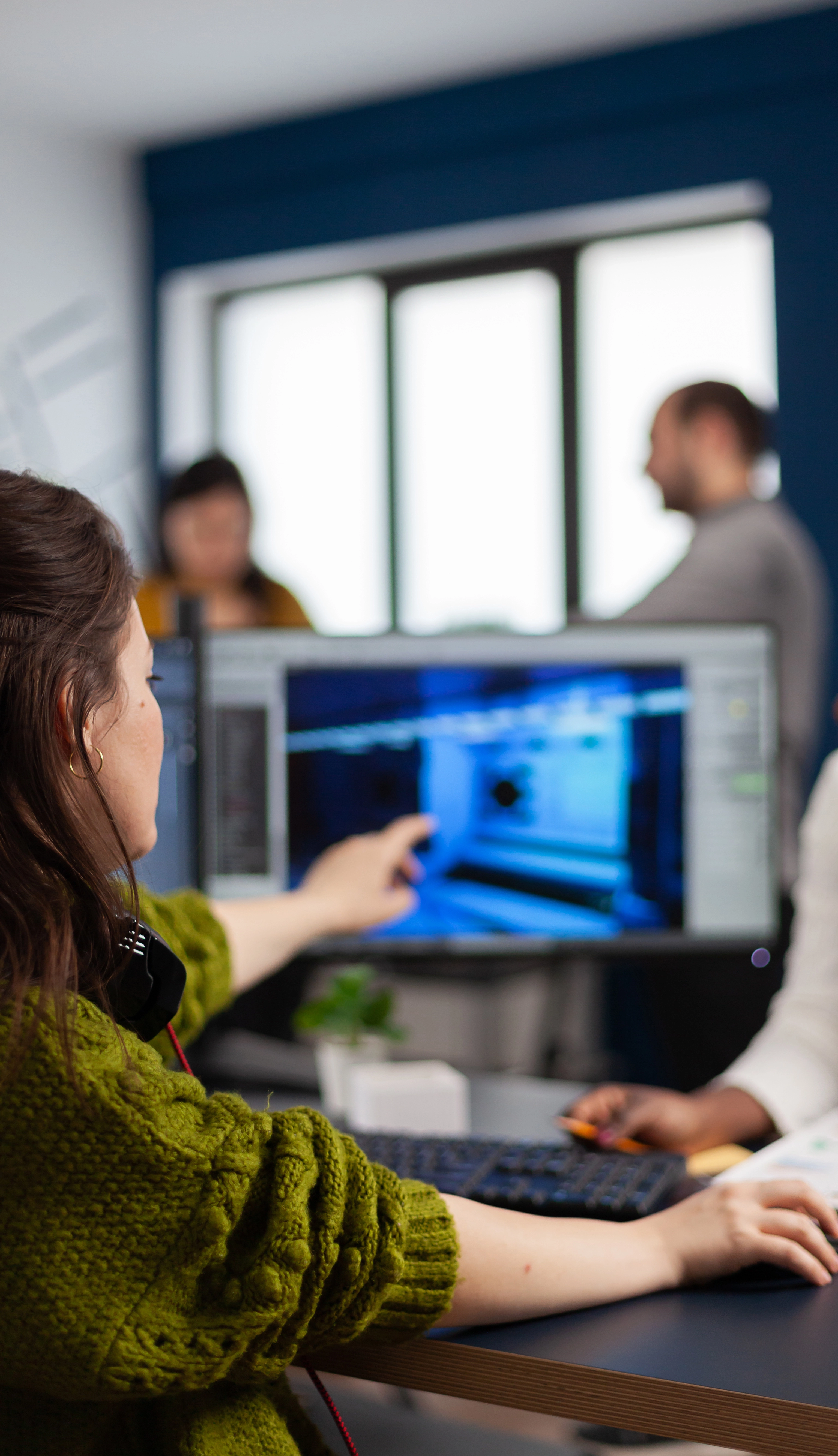 People collaborating in an office, with one person pointing at a computer screen displaying graphics. Other team members are interacting in the background.